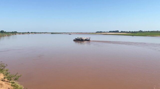 A truck is parked on a wooden ferry while people move around on the riverbank. The sun is shining bright above as they prepare for travel across the water.