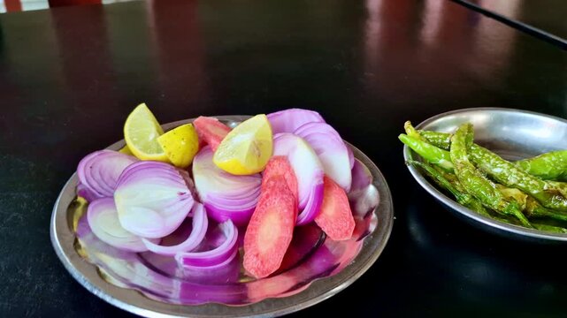 Indian thali side salad in restaurant with sliced onion, lemon, carrot and fried green chillies served with dal in steel bowl on dining table.