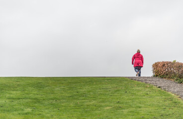 Fototapeta premium Person in red jacket walking up grassy hill under overcast sky symbolizing solitude and journey.