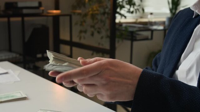 Close-up view of hands of unrecognisable successful businessman wearing white shirt and dark blue jacket holding large stack of dollar bills when sitting at white desk