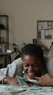 Vertical shot of enthusiastic Black business girl grabbing money after lowering chest on office desk