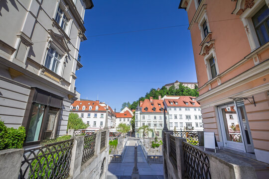 Panoramic view of Ljubljanica river from Ribja brv, also known as the Fish Market Bridge, in the historic city center of Ljubljana, Slovenia, with traditional buildings and red roofs under blue sky.