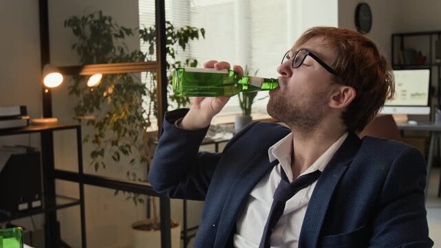 Zoom in rack focus shot of scruffy gen Z man drinking beer and typing on laptop while working at cluttered office desk
