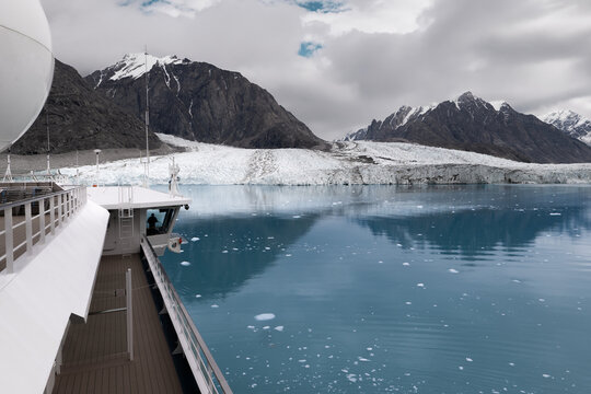 View of a massive glacier flowing into the blue sea with rugged snow-capped mountains under a cloudy sky as seen from a cruise ship Greenland.