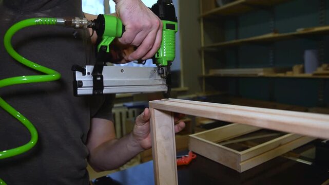 Craftsman in a carpentry workshop hammers nails with a pneumatic hammer. Making a wooden box. A close-up pneumatic nail gun putting nails in boards.