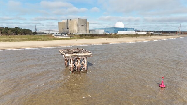 Aerial view of the weathered, skeletal remains of a pier stand starkly against the Sizewell Power Station backdrop, where the sea meets the shore, Leiston, United Kingdom.