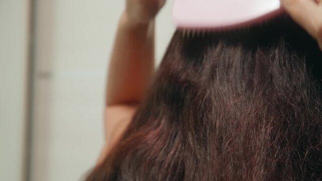 Caucasian woman brushing long brown hair in bathroom mirror smoothing frizz and detangling strands with paddle brush closeup of textured wet hair warm lighting home grooming routine