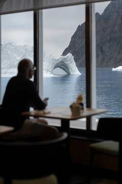 View of a person sitting at a table looking through a window at a large iceberg with a natural arch and dark mountains in Greenland.