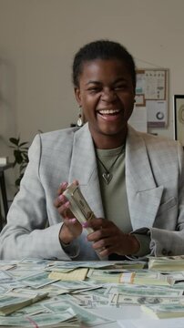 Vertical portrait of excited African American gen Z business girl in office wear smiling at camera smelling freshly printed money