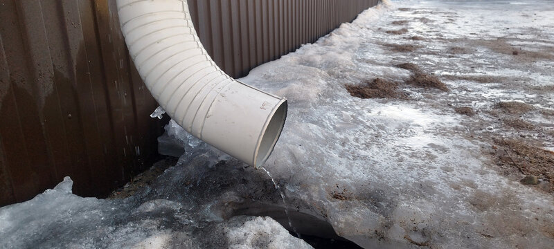 Water trickles from a white plastic downspout onto a patch of melting, dirty snow and ice. The corrugated metal wall of a building stands in the background, signaling the arrival of a spring thaw