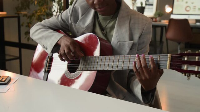 Midsection shot of Black girl in office wear practising playing acoustic guitar at workplace suffering from procrastination