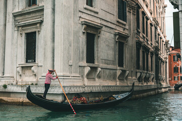 Traditional Venetian gondolier in a red and white striped shirt navigating a black gondola past a grand white historic building, Venice, Italy. © Maria Sap