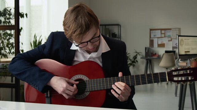 Caucasian gen Z male manager dressed formally and wearing glasses practising playing guitar at workplace