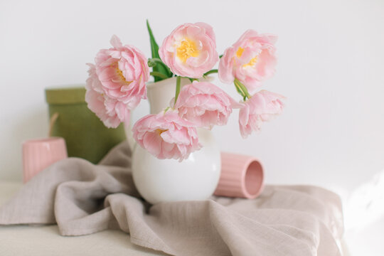 Close-up of Pink peony tulips (double tulips) in a white jug on a table with two ribbed mugs and a linen tablecloth