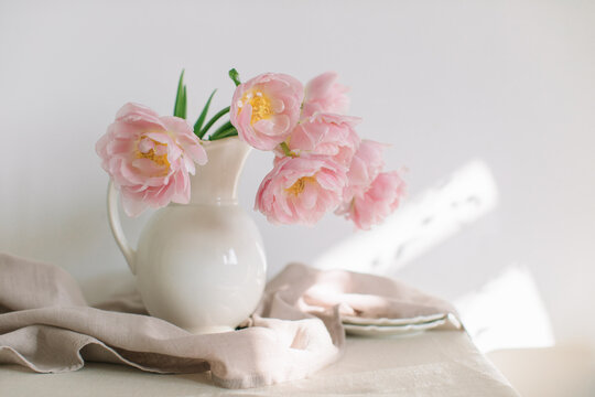 Pink peony tulips (double tulips) in a white jug on a table with two plates and a linen tablecloth in sunlight