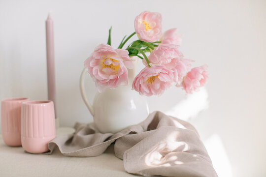 Pink peony tulips (double tulips) in a white jug on a table with a candle, two ribbed mugs and a linen napkin on a sunny day