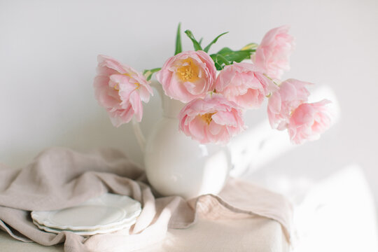 Pink peony tulips (double tulips) in a white jug on a table with two plates and a linen tablecloth in sunlight