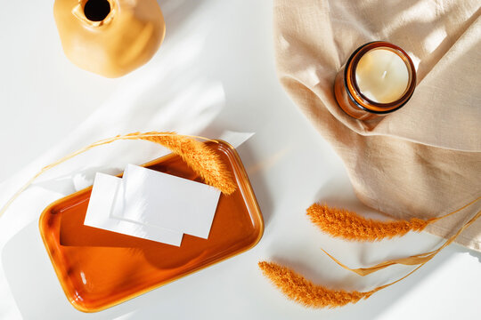Overhead view of two blank white business cards mockup on a glossy orange ceramic tray next to a candle and dried pampas grass on a white table with a linen napkin.