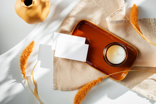 Overhead view of two blank business cards on a ceramic tray next to a scented candle, jug and dried pampas grass with a folded linen napkin on a table