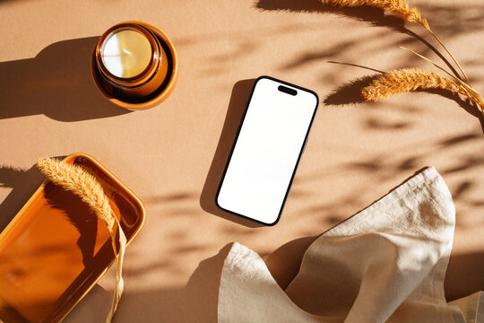 Overhead view of a mobile phone with a blank screen on a table with a ceramic tray, scented candle, napkin and dried pampas grass