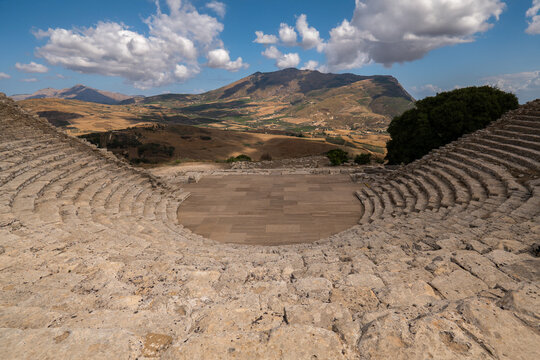 View of the ancient Greek theater of Segesta with its stone tiers overlooking the rolling hills and mountains under a cloudy sky in Segesta, Sicilia, Italy.