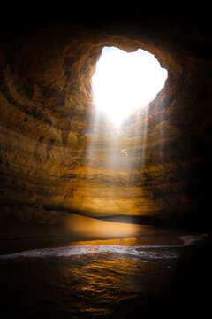 View of the sunlight streaming through the ceiling of Benagil Cave onto the sandy beach and ocean waves in Portimao, Faro District, Portugal.