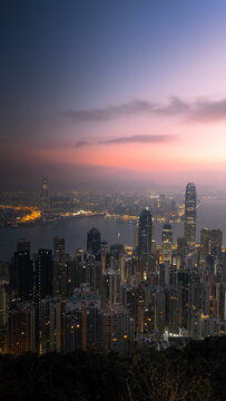 View of the illuminated skyline and Victoria Harbour with skyscrapers under a purple sunset sky in Hong Kong.