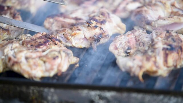 Chicken meat on grill and smoke rises carried with light breeze, boneless drumsticks grilling, man turns pieces with tongs, hot fat sizzling