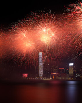 View of vibrant red and orange fireworks exploding over the Victoria Harbour skyline and the International Commerce Centre at night in Hong Kong.