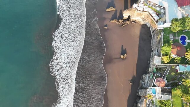 Aerial top-down view of ocean waves rolling onto sandy beach next to luxury villas and tropical coastline, scenic travel destination with clear water and shoreline pattern