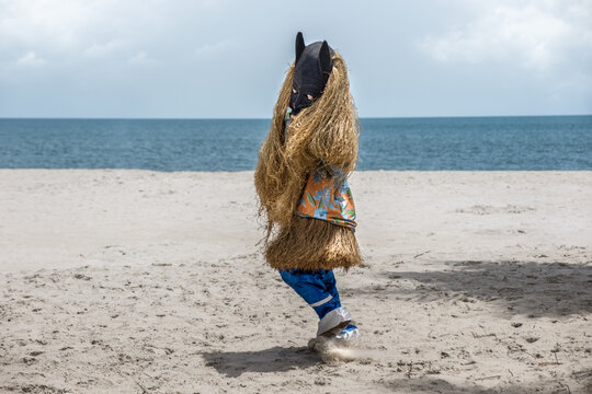 View of a person in a traditional West African masquerade costume with a black mask and raffia fringe dancing on a sandy beach by the ocean in Freetown, Western Area, Sierra Leone.