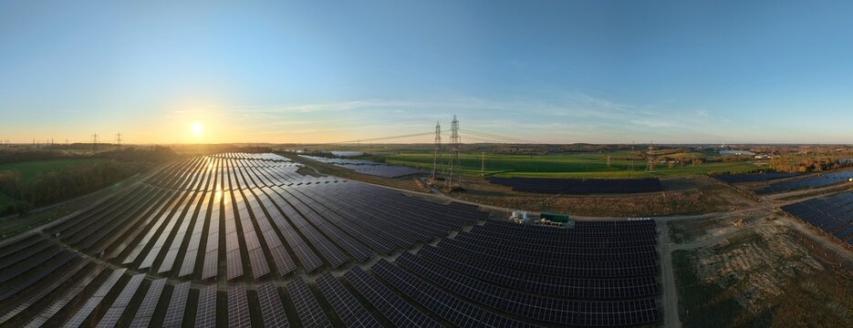 Aerial view of solar panels reflecting the golden sunrise, contrasting with the green fields and distant power lines, Ipswich, United Kingdom.