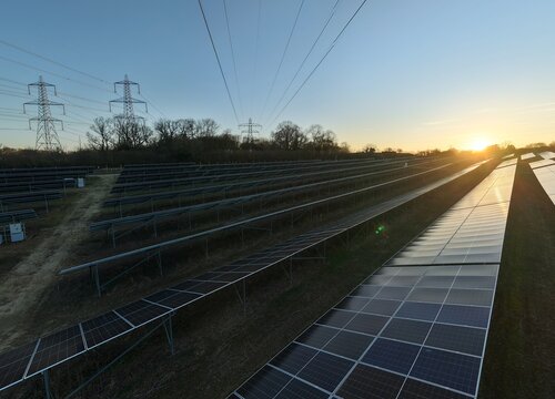 Aerial view of solar panels basking in the golden glow of the setting sun, with electricity pylons standing guard over Tye Lane Solar Farm, Ipswich, United Kingdom.