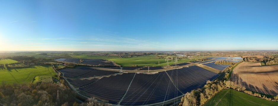 Aerial view of solar panels reflecting the sky's hue, contrasting with the surrounding green and brown fields of Tye Lane Solar farm, Ipswich, United Kingdom.