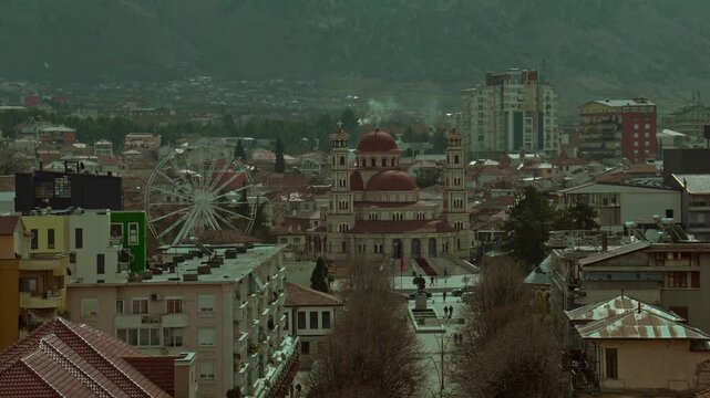 A cinematic aerial view of the Kor&ccedil;&euml; city center in Albania, featuring the Resurrection of Christ Cathedral, the pedestrian square, and a ferris wheel against a mountainous backdrop.
