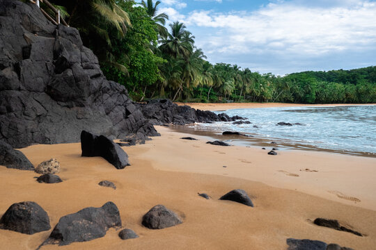 View of a tropical beach with dark volcanic rocks, golden sand, and lush palm trees under a cloudy sky in Sao Tome and Principe.