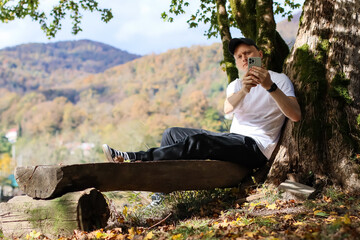 Fototapeta premium Relaxed individual holding smartphone at angle for self-shot on mossy log in fall scenery