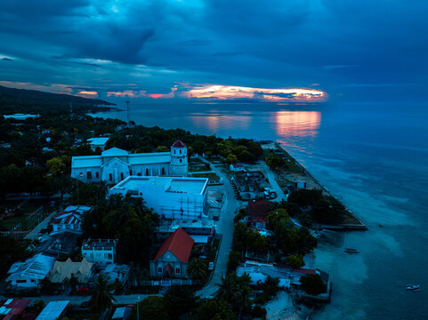 Aerial view of the historic Immaculate Conception Parish Church standing proudly against the dramatic sunset sky, its white facade contrasting with the deep blue sea, Oslob, Cebu, Philippines.