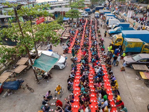 Dhaka, Bangladesh - 17 March 2026: Aerial view of rows of people seated at long tables covered with red cloths, sharing a meal in the bustling city streets.