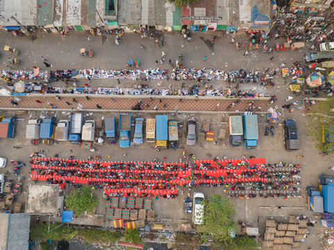 Dhaka, Bangladesh - 17 March 2026: Aerial view of a bustling city scene where rows of trucks line up beside a vibrant gathering of people seated on red mats.