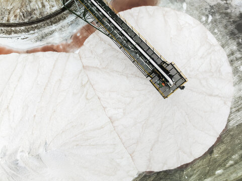 Aerial view of the stark white salt piles meeting the rose-tinted brine ponds under the watchful gaze of a metal conveyor, Walvis Bay, Erongo Region, Namibia.