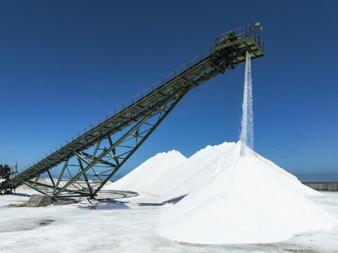 Aerial view of gleaming white salt piles cascade from a green conveyor against the stark blue sky at the saltworks., Walvis Bay, Erongo Region, Namibia.