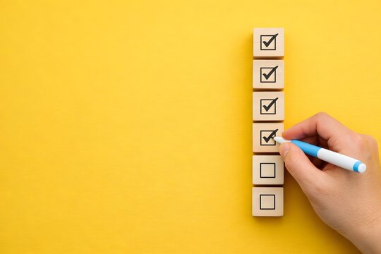 Hand with a blue pen is marking check boxes on a stack of wooden blocks against a bright yellow background, representing completion and achievement.