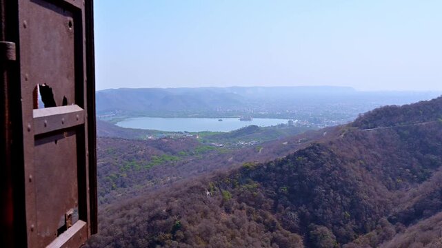 Panoramic view from Jaigarh Fort Jaipur overlooking Aravalli hills and Man Sagar Lake with Jal Mahal at center, seen through a rustic fort window.