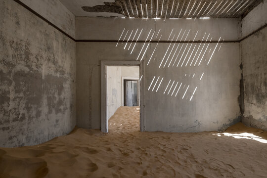 View of the interior of an abandoned house filled with desert sand dunes and sunlight filtering through a broken wooden ceiling in Kolmanskop, Karas Region, Namibia.