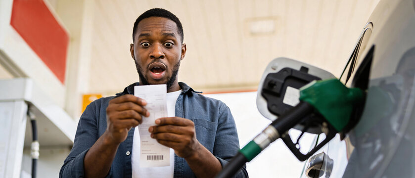 Soaring petroleum prices, energy market surge oil crisis, African American male driver shocked with paper receipt at gas station with gasoline fuel nozzle