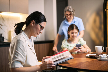 Fototapeta premium Young adult mother reading at table while teenage daughter using smartphone and senior woman standing nearby. Suitable for family finance, parenting, digital habits, multigenerational home life