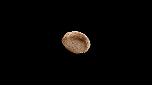 Close-up of a single, small, golden-brown bread roll rotating slowly against a stark black background, showcasing its texture and form.