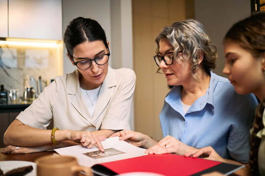 Young adult woman guiding middle aged woman and girl while reviewing family photo album at table. Suitable for family history, caregiving support, memory preservation marketing, medium shot