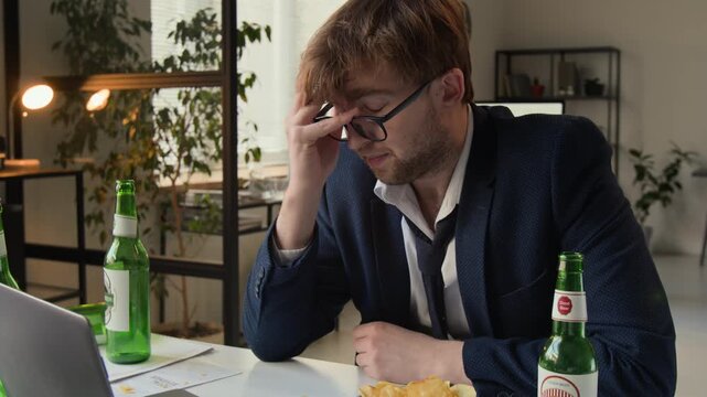Tilt up view of exhausted gen Z man removing glasses and rubbing his eyes resisting burnout while sitting at messy desk featuring several empty beer bottles